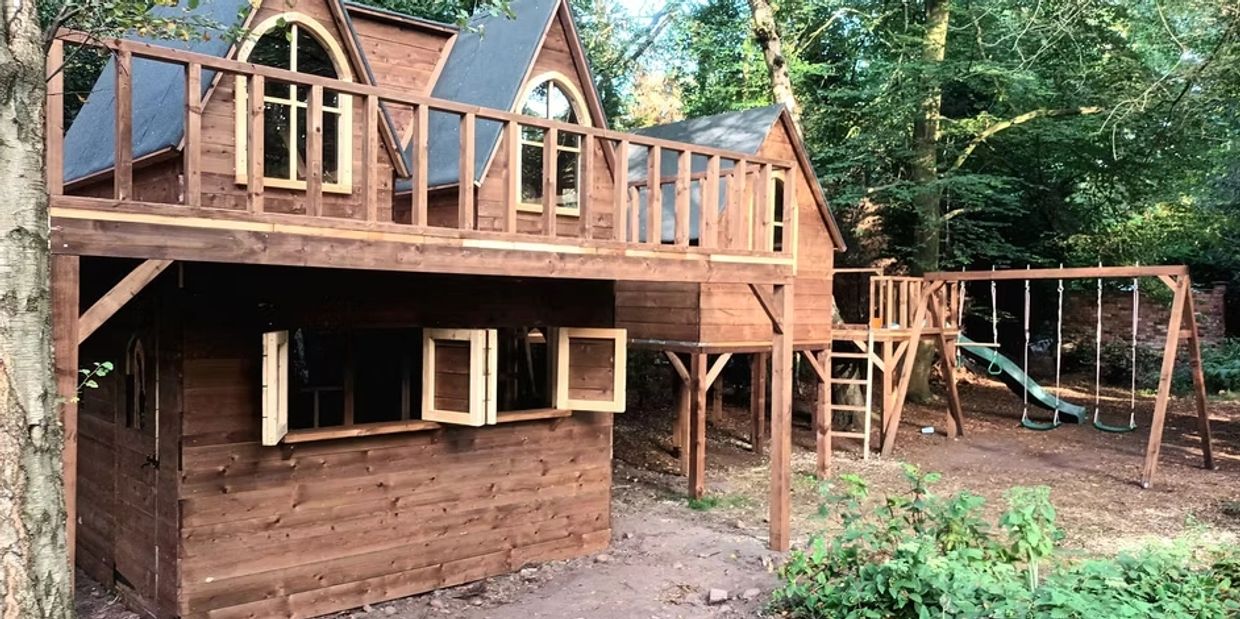 A wooden playhouse with swings and a slide in a forested backyard.