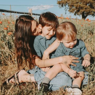 A mother hugs and kisses her two young children outdoors.