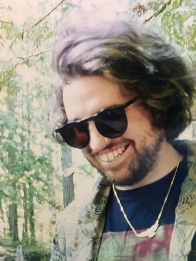 A smiling man with sunglasses and a beard in an outdoor forest setting.