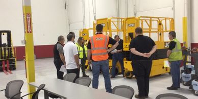 Group of workers in a warehouse listening to instructions near yellow machinery.