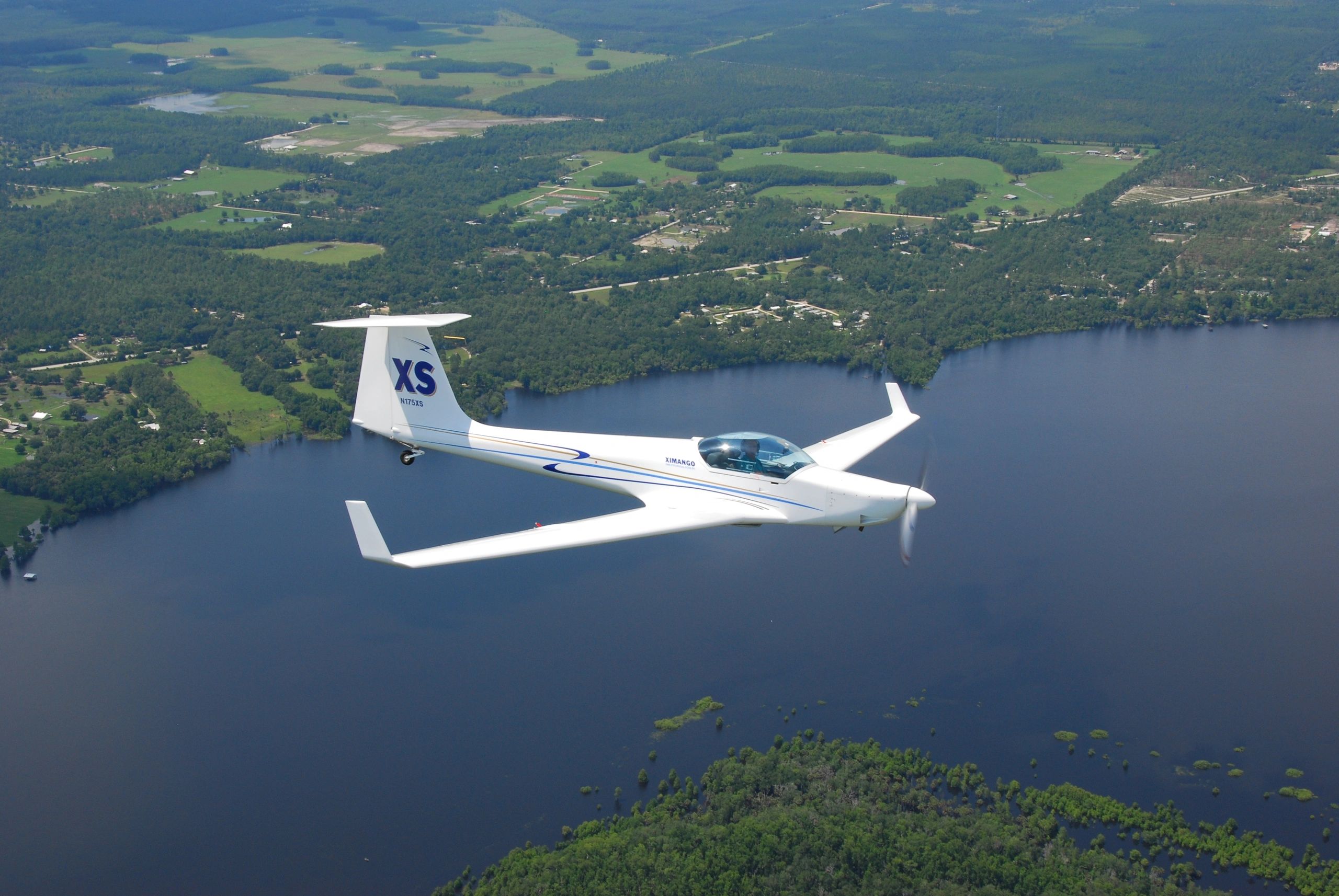 A white glider plane flying over a lake and forest landscape.