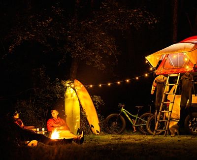 Two people camping at night with a campfire, bicycles, and a rooftop tent lit by string lights.