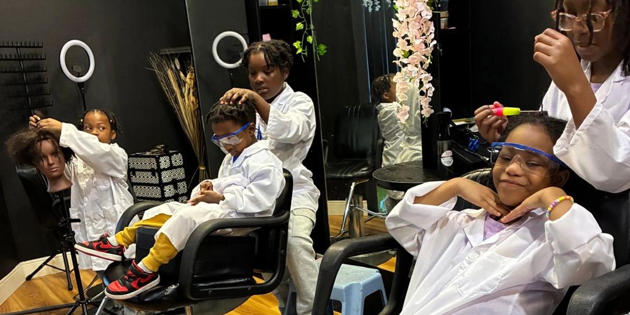 Children role-playing as hairstylists in a salon setting with mannequin heads and safety glasses.