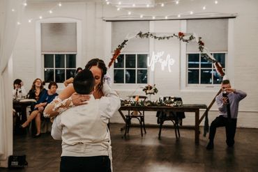 A couple shares a dance at their wedding with guests watching and a photographer capturing the moment.