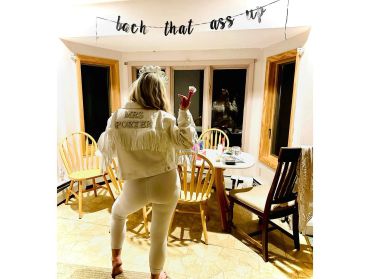 Bride-to-be wearing a custom jacket stands in a cozy dining area with a celebratory banner.