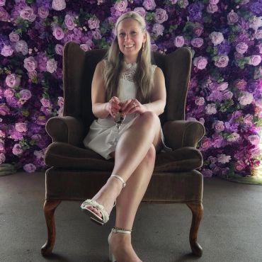 Woman in white dress sitting on a brown armchair with a floral purple backdrop.