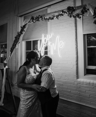 Two brides share a kiss beneath a glowing 'You + Me' sign at their wedding.