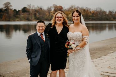 Three people smiling by a lakeside, including a bride in a wedding dress.
