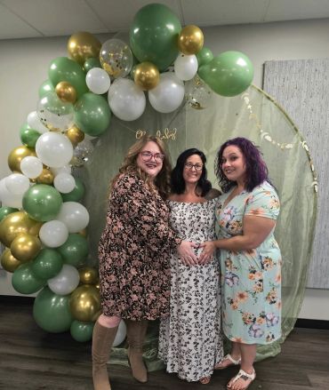 Three women posing at a baby shower with balloon decor.