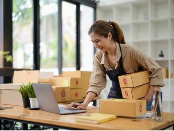 Mujer revisando laptop mientras sostiene cajas.