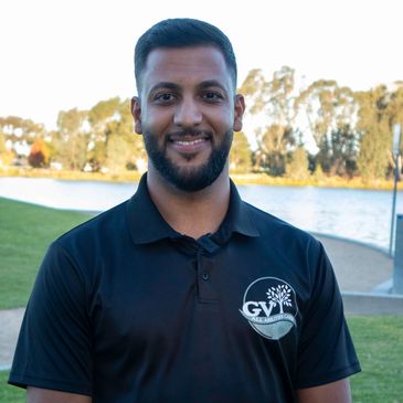 Smiling man in a black polo shirt by a lakeside park.
