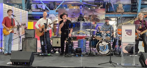 A band performs on stage with a helicopter backdrop in an industrial setting.