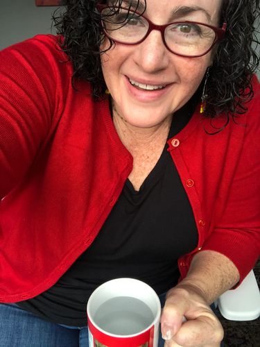 Smiling therapist in purple glasses, jeans, black shirt and red cardigan smiles holding a red mug.