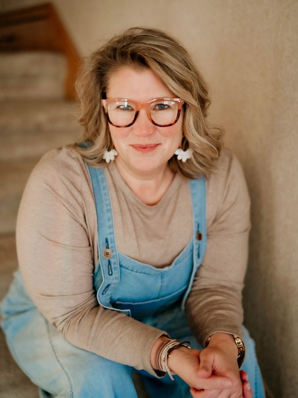 Smiling woman with glasses wearing a beige shirt and denim overalls.