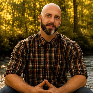 Man with a beard in a plaid shirt sitting outdoors with a forest background.