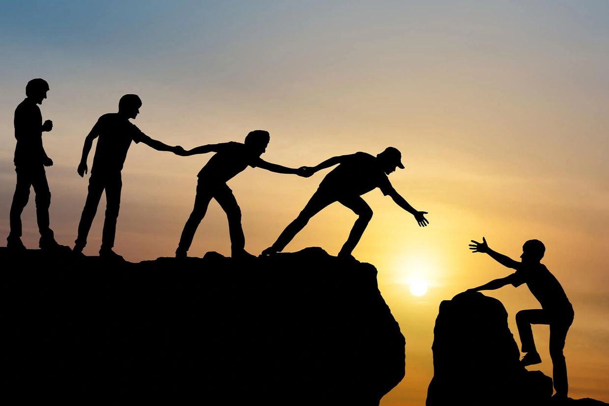 Silhouettes helping each other climb a steep cliff at sunset.