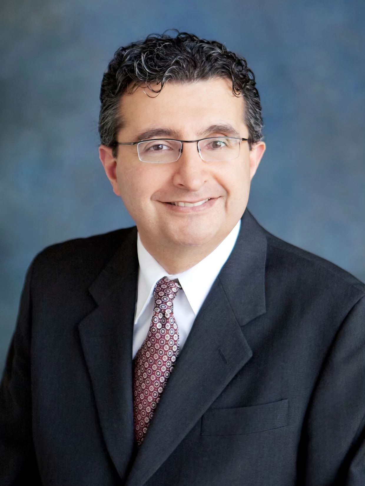 Professional man in suit and glasses smiling against a blue background.