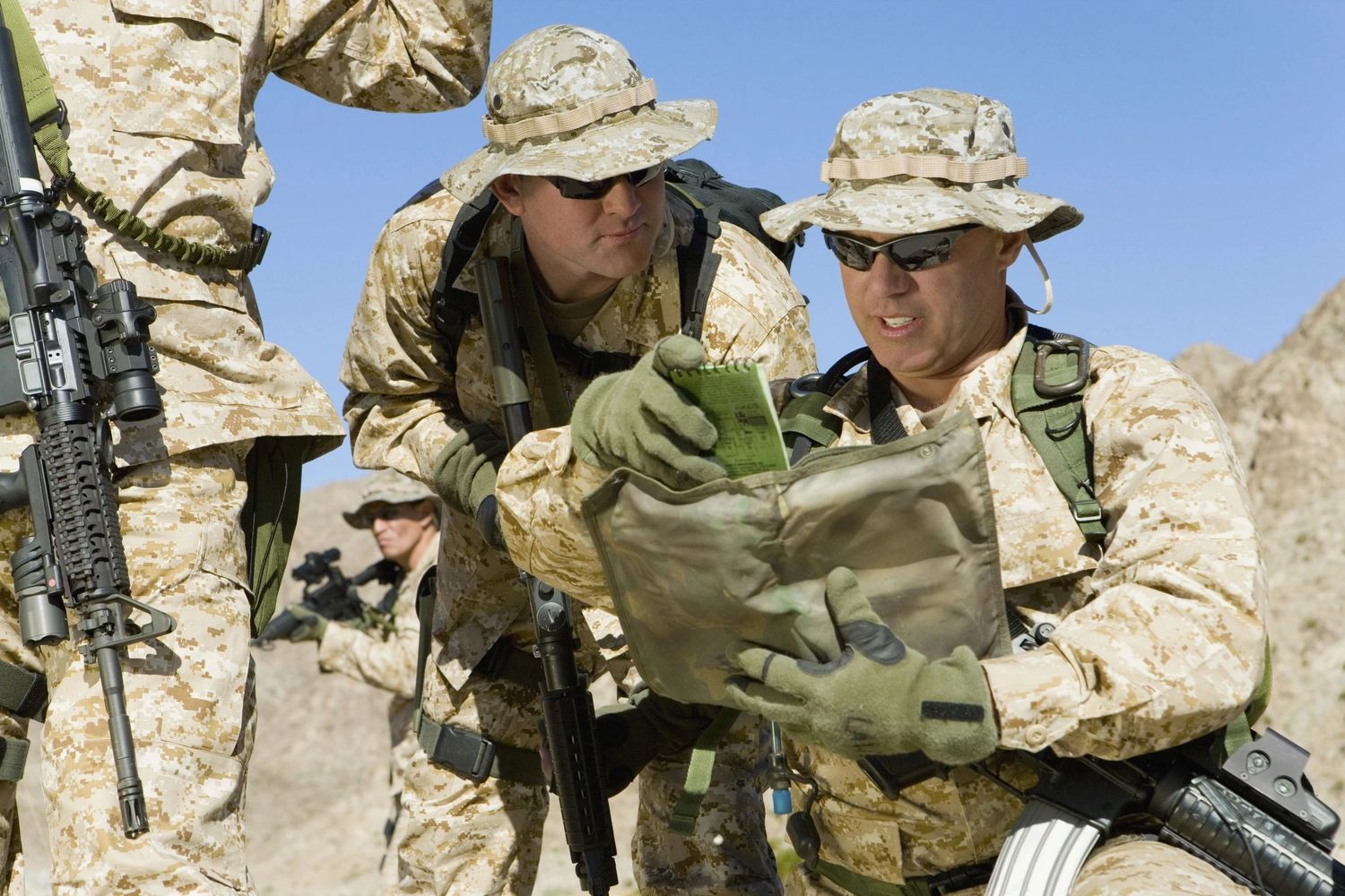 Soldiers in desert camouflage gear examining a map in a rocky terrain.
