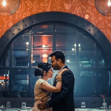 A couple embraces tenderly under a large arched window in a warmly lit room.
