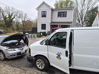 Person checks engine of a silver car by a white van in front of a two-story house.
