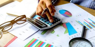 Person using calculator with financial charts and glasses on desk.