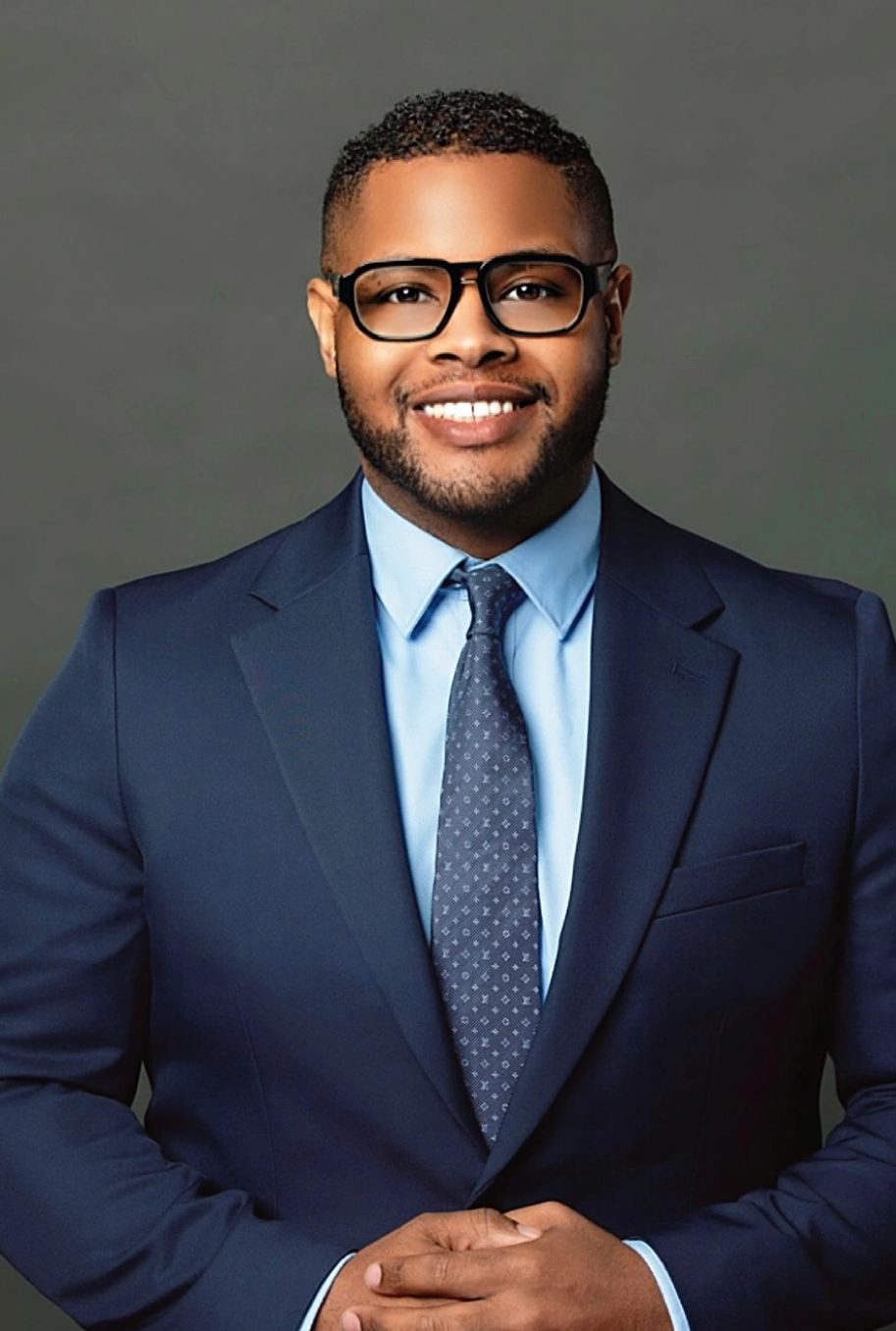 Confident man in a navy suit and glasses smiling.