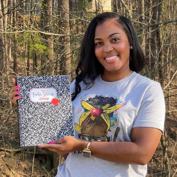 A smiling woman holds a journal titled 'The Teacher Self-Care Journal' outdoors.