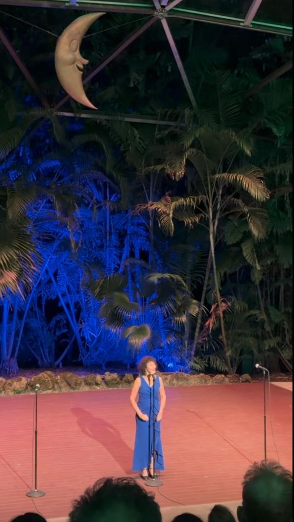 Woman in a blue dress performing on stage with tropical backdrop.