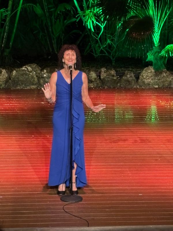 Woman in blue dress singing on stage with green plants behind.