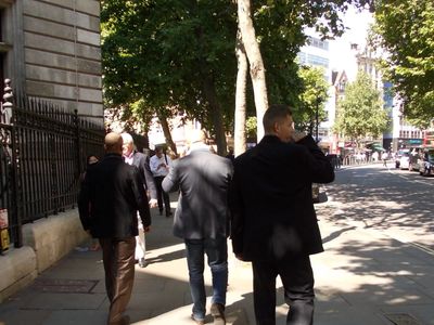 People walking on a sunny city sidewalk with trees and cars nearby.