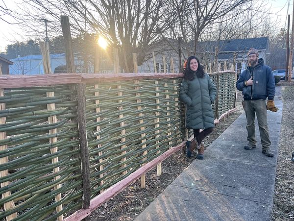 Two people standing by a woven wooden fence at sunset.