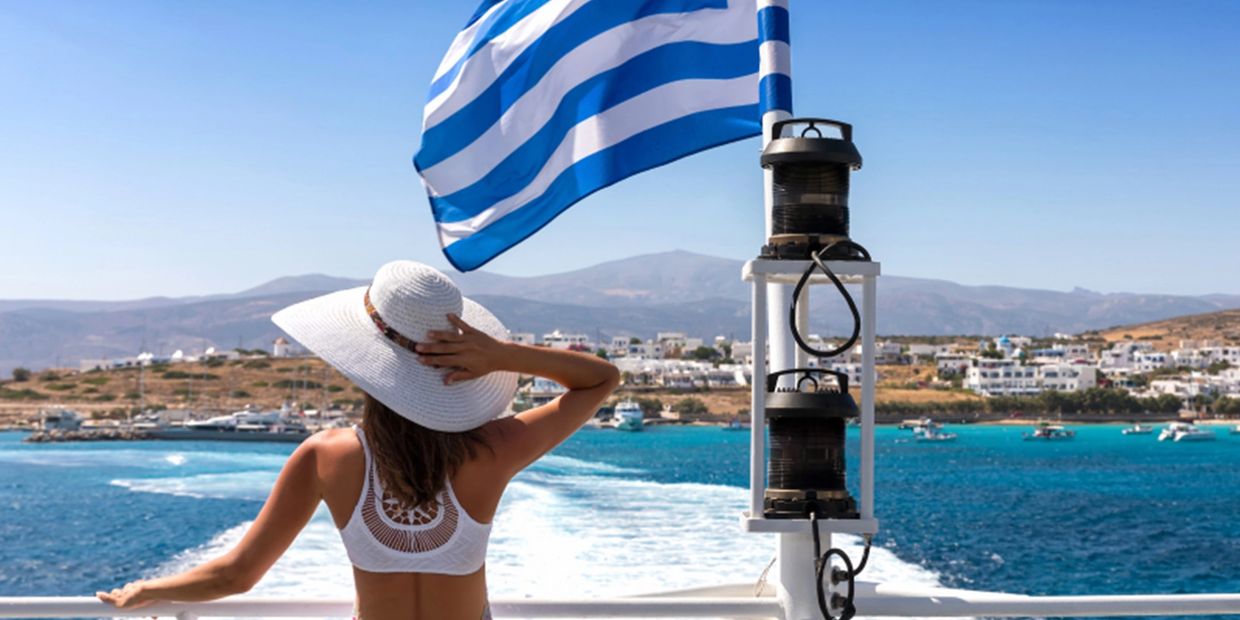Woman in white hat on boat with Greek flag and coastline.