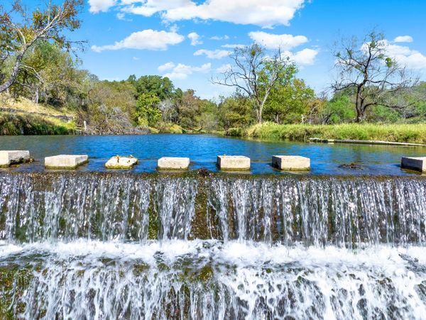 Water cascades over a small dam with stepping stones and lush greenery under a bright blue sky.