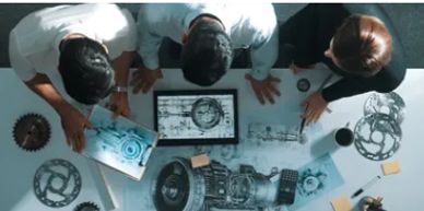 Three people examining mechanical blueprints around a table with gears and parts.