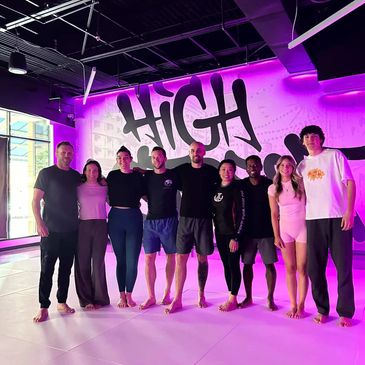 Group of eight people posing barefoot in front of a 'HIGH' sign with purple lighting.