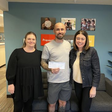 Three people standing indoors, smiling, with one holding a check.