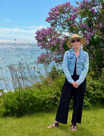 Woman in hat and denim jacket by water and blooming lilac bush.