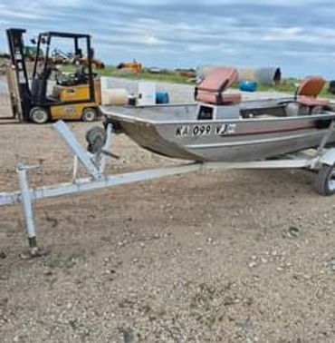 Small aluminum boat on a trailer in a gravel yard with a forklift in the background.