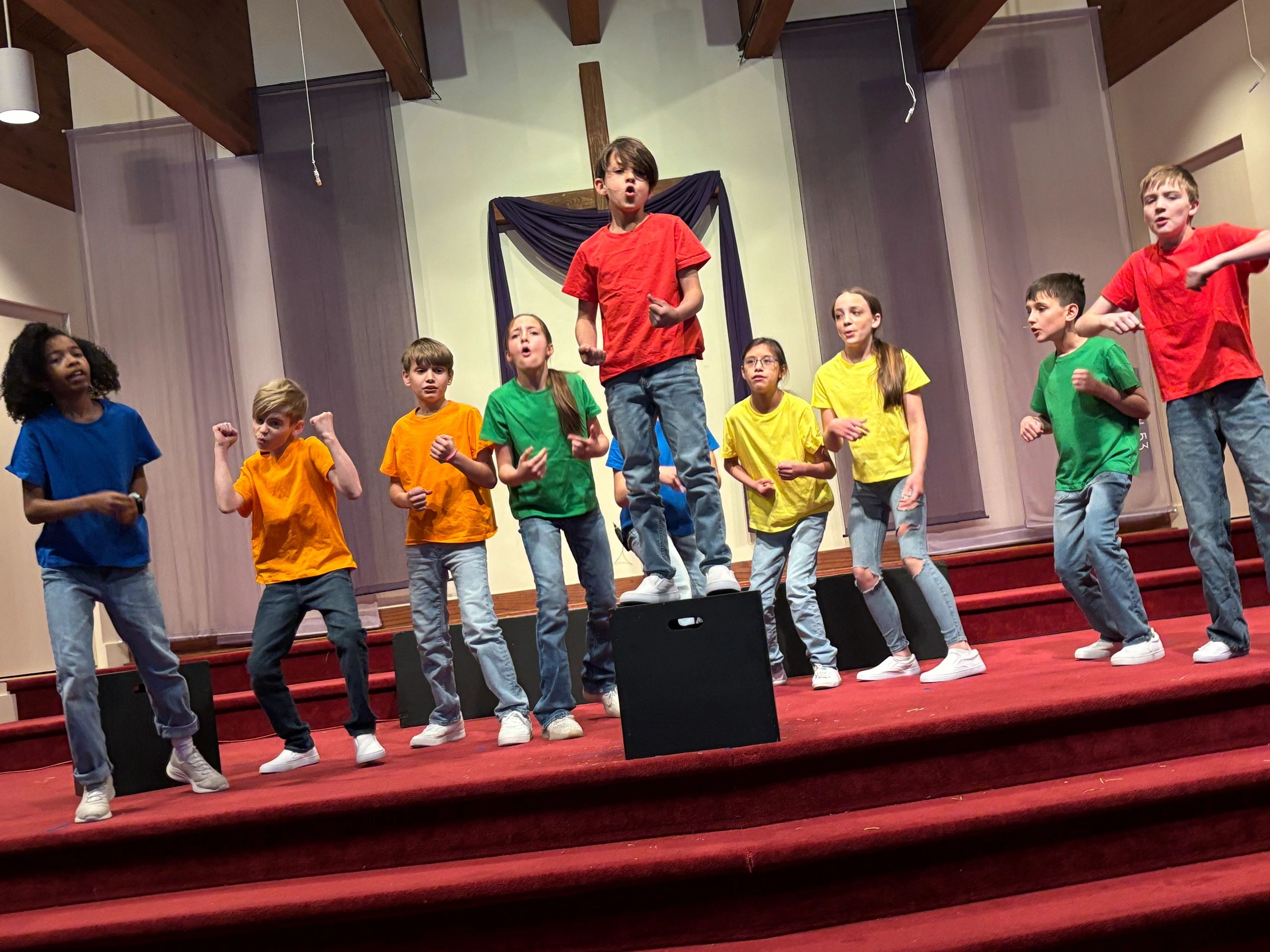 Kids in colorful shirts performing energetically on stage with a cross backdrop.