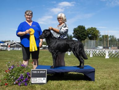 Two women with a black dog winning a third place ribbon at a dog show.