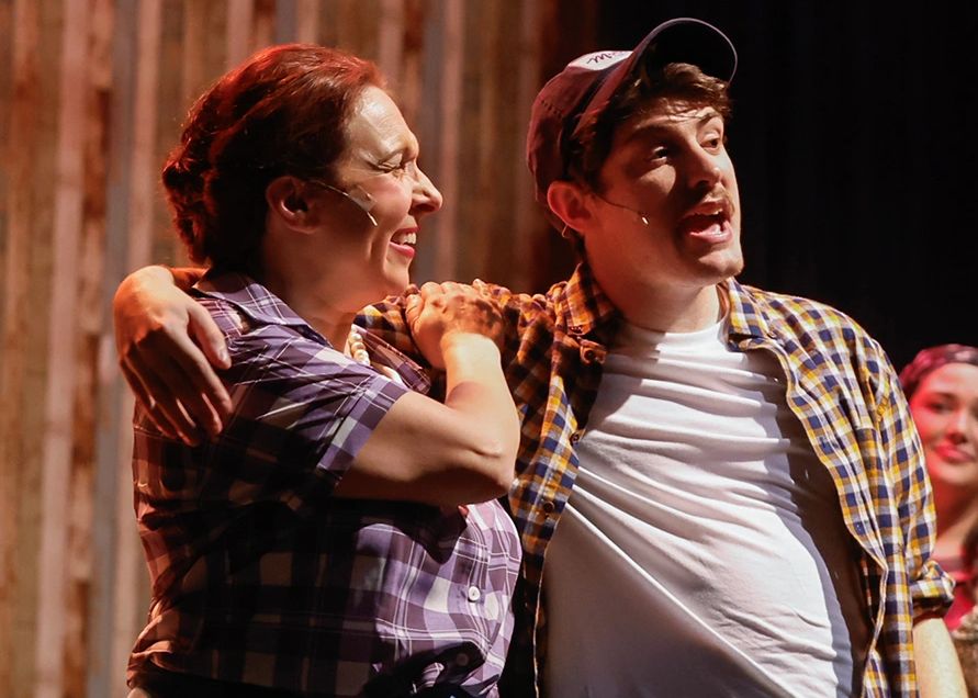 Two actors in casual plaid shirts performing on stage with microphones.