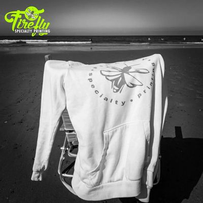 White hoodie with Firefly Specialty Printing logo draped over a chair on the beach.