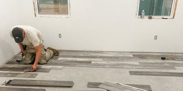 Worker installing gray wooden floor planks in a room with white walls.