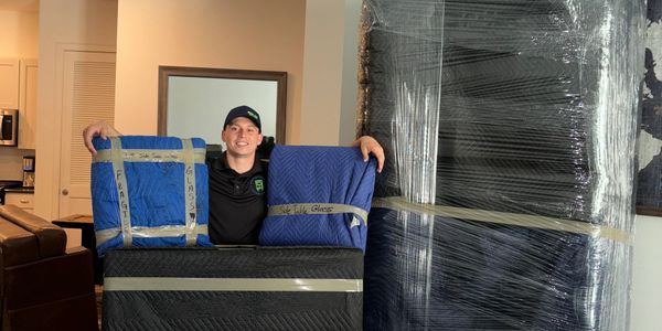 Smiling mover posing with wrapped furniture in a cozy living room.