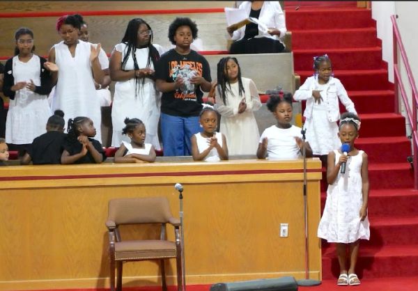 Young children performing in a church choir, one girl sings with a microphone.