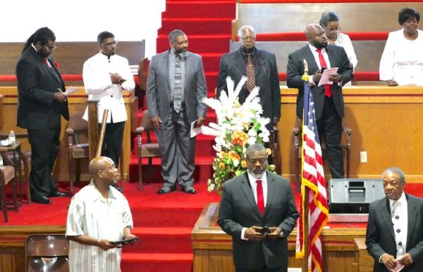 Men in suits and formal wear gathered in a church setting with an American flag and floral arrangement.