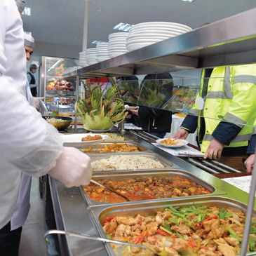 People serving and receiving food at a cafeteria counter with various dishes.