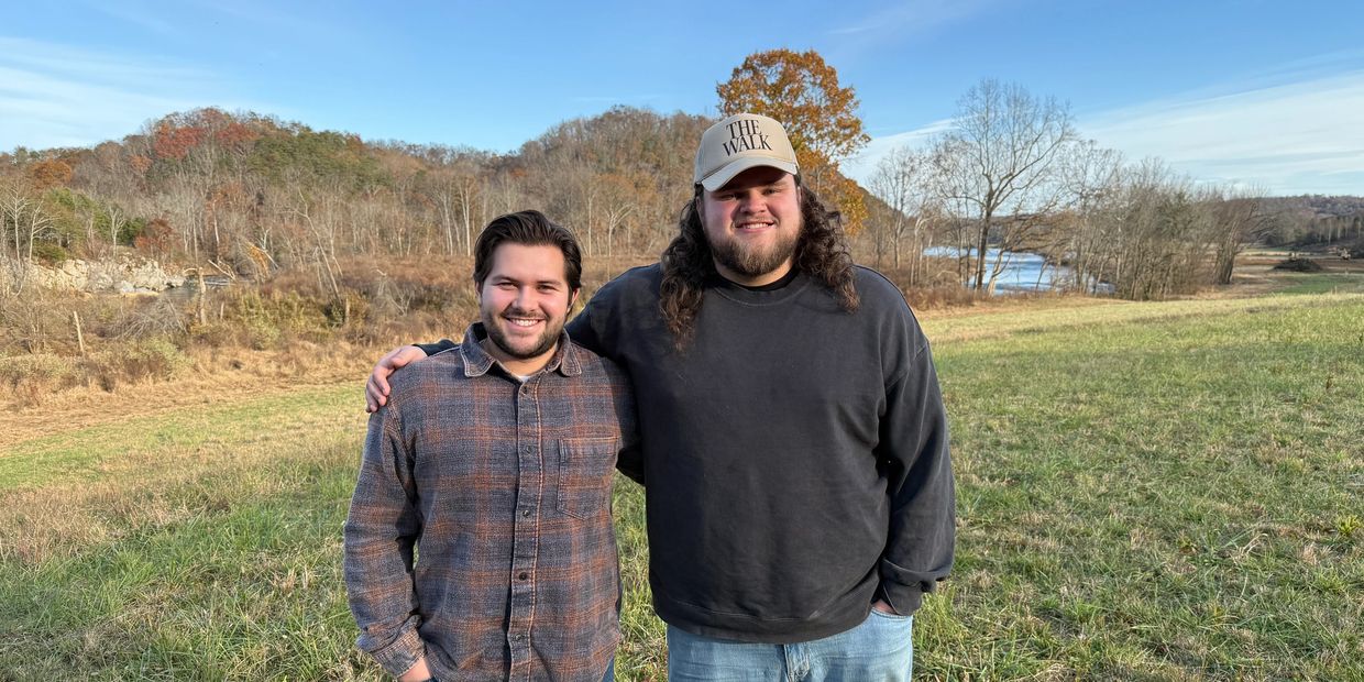 Two men smiling and standing together in a grassy field with trees and hills in the background.
