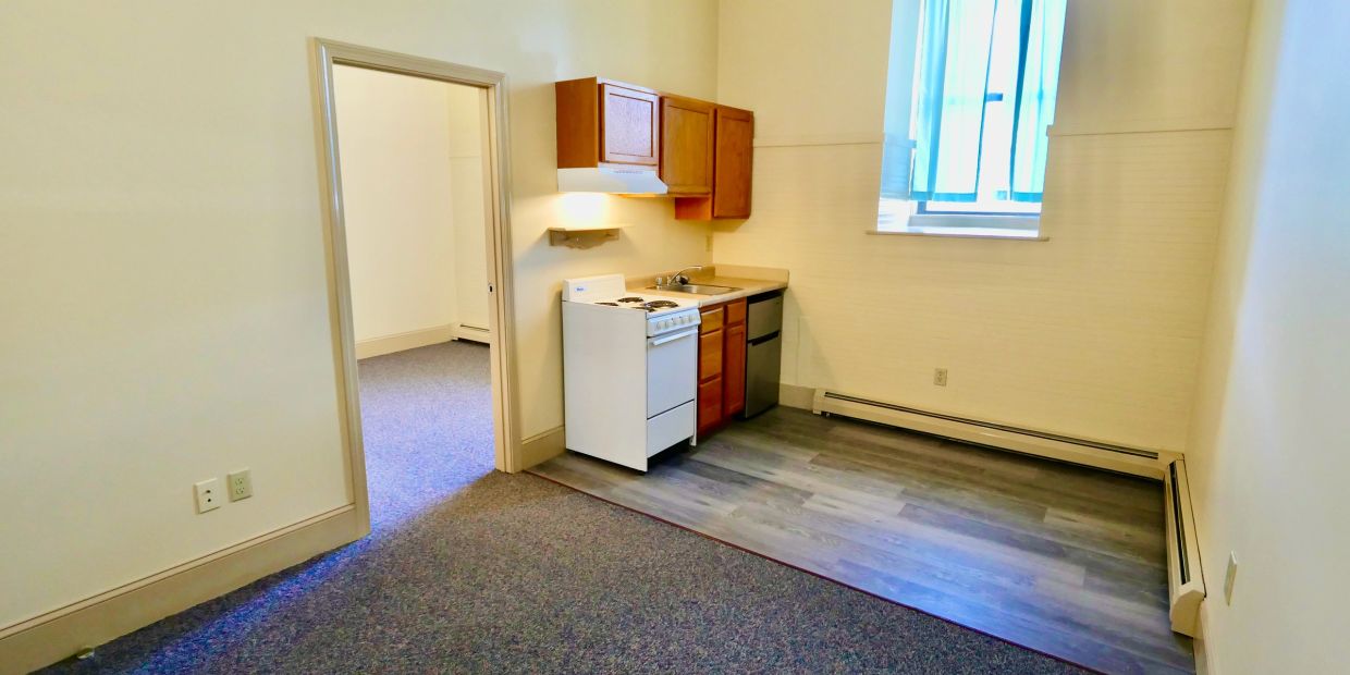 Small kitchen area with stove, sink, and cabinets in an empty room.
