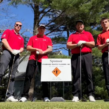 Four men in red shirts posing on a golf course behind a sponsor sign.